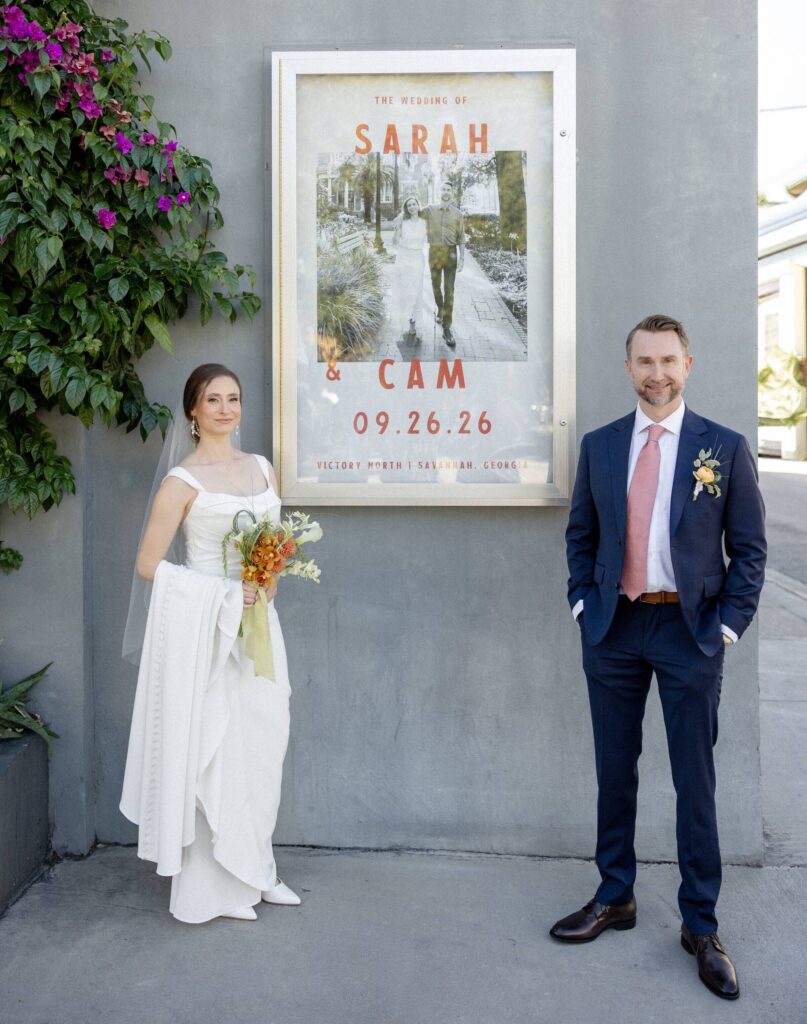 Cam and Sarah standing on either side of a wedding poster that reads Sarah and Cam, 09.26.26
